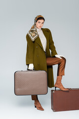 full length of trendy woman in green coat and headscarf posing with retro suitcases on grey.