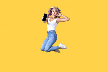 Full length portrait of young Asian woman holding smartphone jumping on yellow background. Cheerful young female jumping up and showing mobile phone with empty screen, in studio