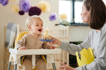 Little kid happy with piece of cake with candle while sitting on chair and celebrating birthday...