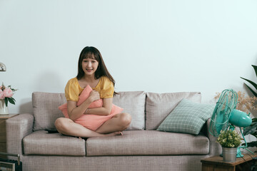 asian lady facing camera is relaxing on sofa at home. japanese woman holding pillow in arms is...