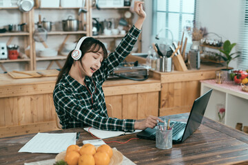 asian female student is relaxing at dining table listening to upbeat songs on laptop. chinese girl...