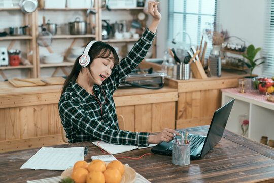 asian female student is relaxing at dining table listening to upbeat songs on laptop. chinese girl taking break from preparing for test is enjoying online streaming music with hand raising in the air