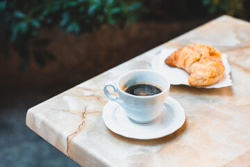 Cup of coffee and a croissant for breakfast on a table in a street cafe.