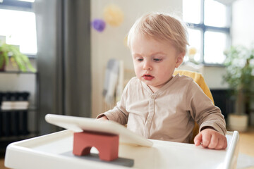 Little child with blond hair sitting on his chair and watching cartoons with interest on digital tablet