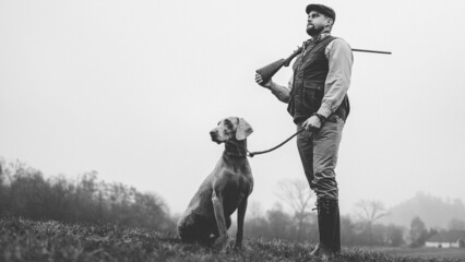 Hunter man with dog in traditional shooting clothes on field holding shotgun, black and white photo.