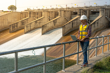 A dam engineering doing his checking routine. He is wearing a white hard hat and yellow transparent vest. He is standing by the rail by the dam.