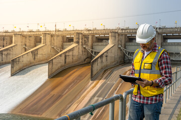 A dam engineering doing his checking routine. He is wearing a white hard hat and yellow transparent vest. He is standing by the rail by the dam.