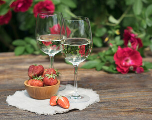 Two glasses with white wine and with a reflection of roses in the glass. Strawberries standing on a table and a linen napkin against a background of a bush of red roses.