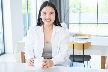  smiling young asian woman in white shirt with cup of tea or coffee and laptop at modern home