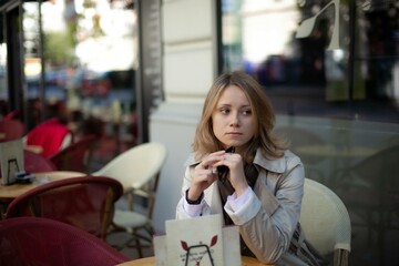 Young beautiful woman holding sunglasses in hand smiling and sitting alone by table of summer cafe