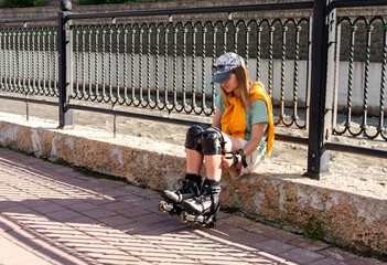 young woman in a yellow sweatshirt cap in protective equipment and roller skates sits on the embankment relaxing after roller skating at summer - obrazy, fototapety, plakaty