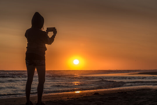 Silhouette of a hoodie woman on the beach taking a photo with her smartphone at sunset with the sun on the horizon.