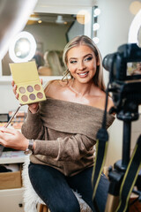 Happy young woman streaming a beauty makeup vlog from home or workshop. Beautiful online content creator cosmetician applying makeup and explaining some work tools. Vlogging and online channel work.