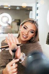 Obraz na płótnie Canvas Happy young woman streaming a beauty makeup vlog from home or workshop. Beautiful online content creator cosmetician applying makeup and explaining some work tools. Vlogging and online channel work.