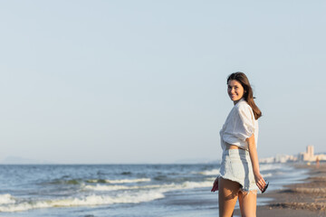 Fototapeta na wymiar Happy young woman holding sunglasses and looking at camera near sea.