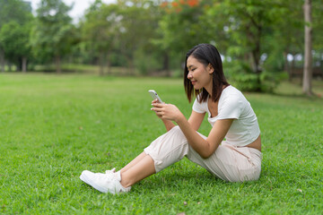 Young beautiful asian woman sitting on the grass and smiling happily while using her phone and enjoying her free time happily in a public park