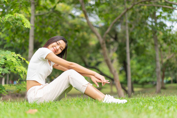 Naklejka na ściany i meble Portrait photo of a young beautiful asian female lady in white top sitting on the grass embracing the breeze wind and soft sunlight relaxingly and chillingly in a public park