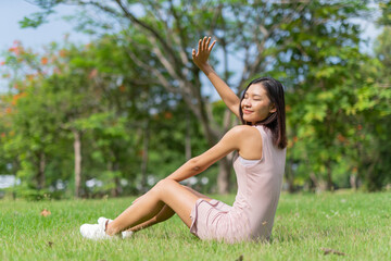 Portrait photo of a young beautiful asian female lady in pink dress sitting on the grass embracing the breeze wind and soft sunlight relaxingly and chillingly in a public park