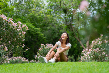 Naklejka na ściany i meble Portrait photo of a young beautiful asian female lady in white top sitting on the grass embracing the breeze wind and soft sunlight relaxingly and chillingly in a public park