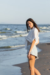 Pretty brunette woman in shirt holding sunglasses and looking at camera on beach.