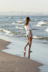 Fototapeta na wymiar Side view of young woman in shorts standing on wet sand near sea.