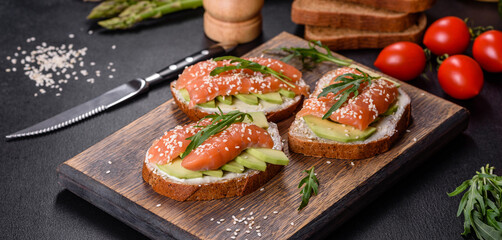 Toast sandwich with butter, avocado and salmon, decorated with arugula and sesame seeds, on a black stone background