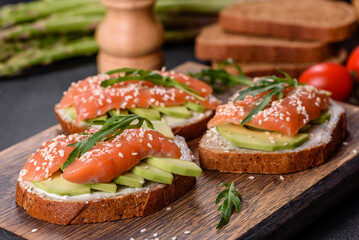 Toast sandwich with butter, avocado and salmon, decorated with arugula and sesame seeds, on a black stone background