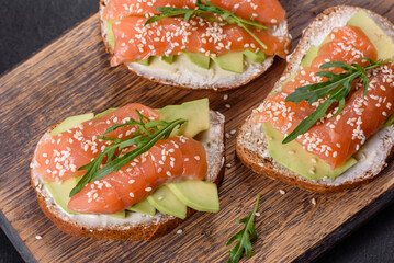 Toast sandwich with butter, avocado and salmon, decorated with arugula and sesame seeds, on a black stone background