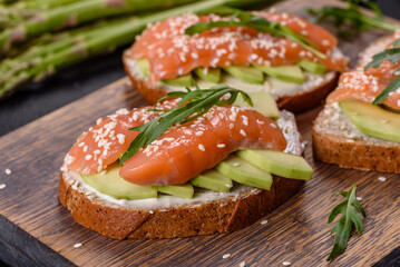 Toast sandwich with butter, avocado and salmon, decorated with arugula and sesame seeds, on a black stone background