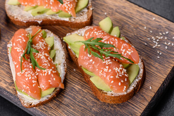 Toast sandwich with butter, avocado and salmon, decorated with arugula and sesame seeds, on a black stone background