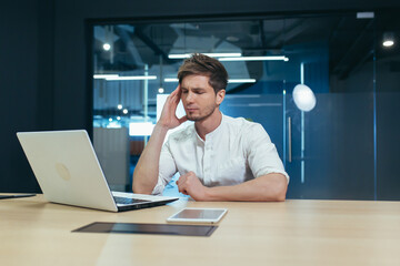 Sick and tired man working in the office with a laptop, has a severe headache, overworked businessman worker