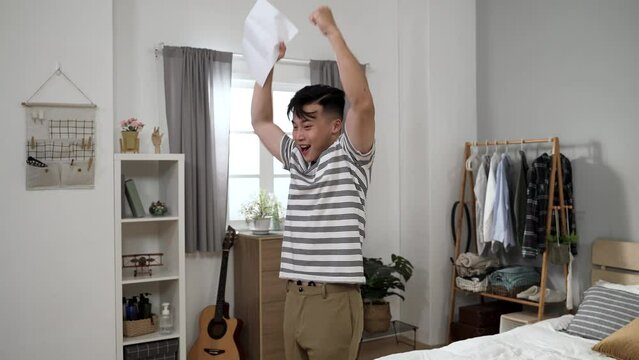 happy asian young man looking at paper document in surprise is kissing it with raised fists while celebrating success of getting admitted to dream school at home