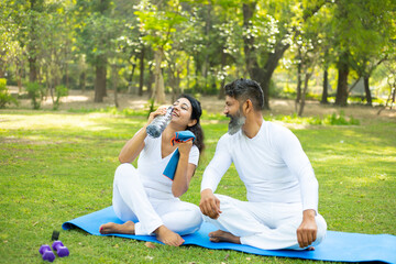 Indian man and woman sitting in the park drinking water after yoga exercise workout, Asian couple relaxing in the garden together outdoor.