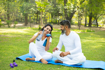 Indian man and woman sitting in the park drinking water after yoga exercise workout, Asian couple relaxing in the garden together outdoor.
