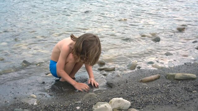 Cute little boy with long blond hair playing with sand nearby the see.