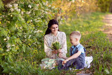 Mom is sitting next to her son on the grass , the boy is holding flowers in his hands