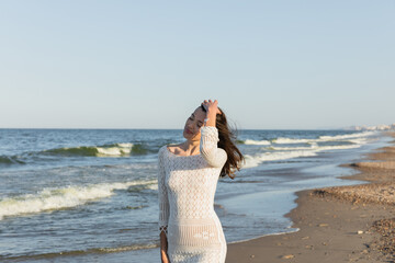 Fototapeta na wymiar Brunette woman in knitted dress touching hair near sea on beach.