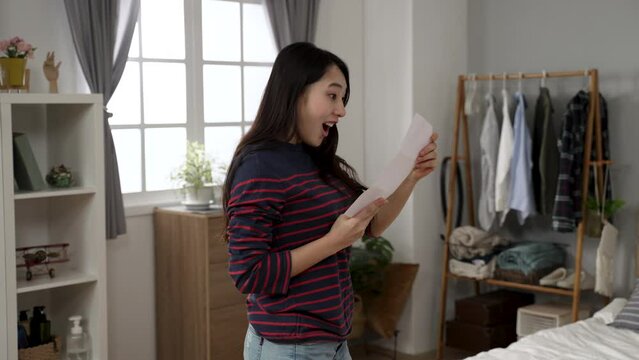 excited Japanese female unfolding letter with job offer message while walking in bedroom is spinning in place with clenched fist and reading details with smile