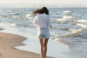 Back view of woman in white shirt and denim shorts walking near sea on beach.