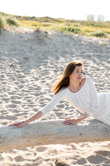 Fototapeta na wymiar Woman in summer white dress posing on wooden log on beach.