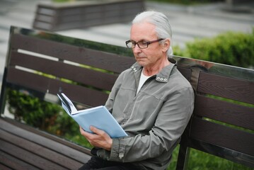 Fototapeta premium Portrait of senior man reading on bench during summer day.