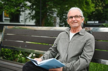 Fototapeta premium Old man with gray hair reads a book on a bench in the park. Rest in the park.