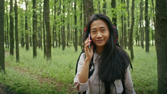 happy asian woman with a backpack looking for a way in the forest, using a phone, talking on a video call, looking at a map. camping in summer.