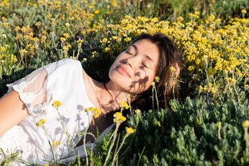 Brunette woman in white dress lying near flowers and grass on meadow.