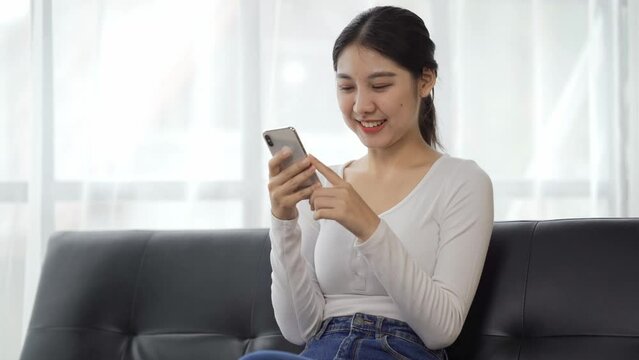 Happy woman relaxing on comfortable couch, holding smartphone in hands. Smiling young lady chatting in social networks, watching funny videos, using mobile applications at home.