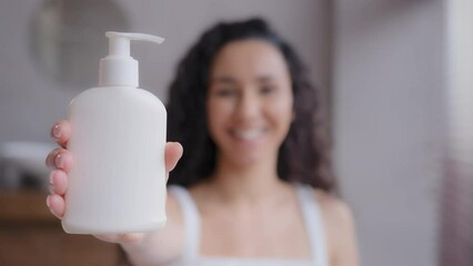 Close-up young attractive happy woman standing in bathroom looking at camera showing moisturizing body lotion in dispenser advertising shower gel skin care cream promotional video out of focus girl