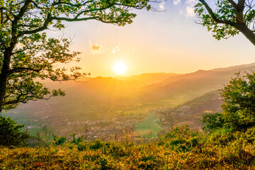 Fototapeta na wymiar wonderful spring sunset in mountains with amazing view from highland among greeen tree branches to a valley with town, mountains and beautiful sunset on background