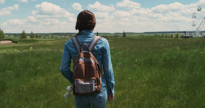 Young woman with backpack dressed in denim walks through green field with tall grass. Rear view, camera follows girl. White clouds blue sky, nature, summertime, daytime.