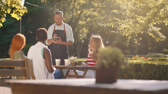 In a forest scenery, a very attractive waiter with a beard and hair tied back is taking orders from a table with three beautiful women
