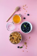 Homemade granola with nuts and seeds in jar, honey and blueberries on pink background. Granola for healthy breakfast. Top view, copy space.